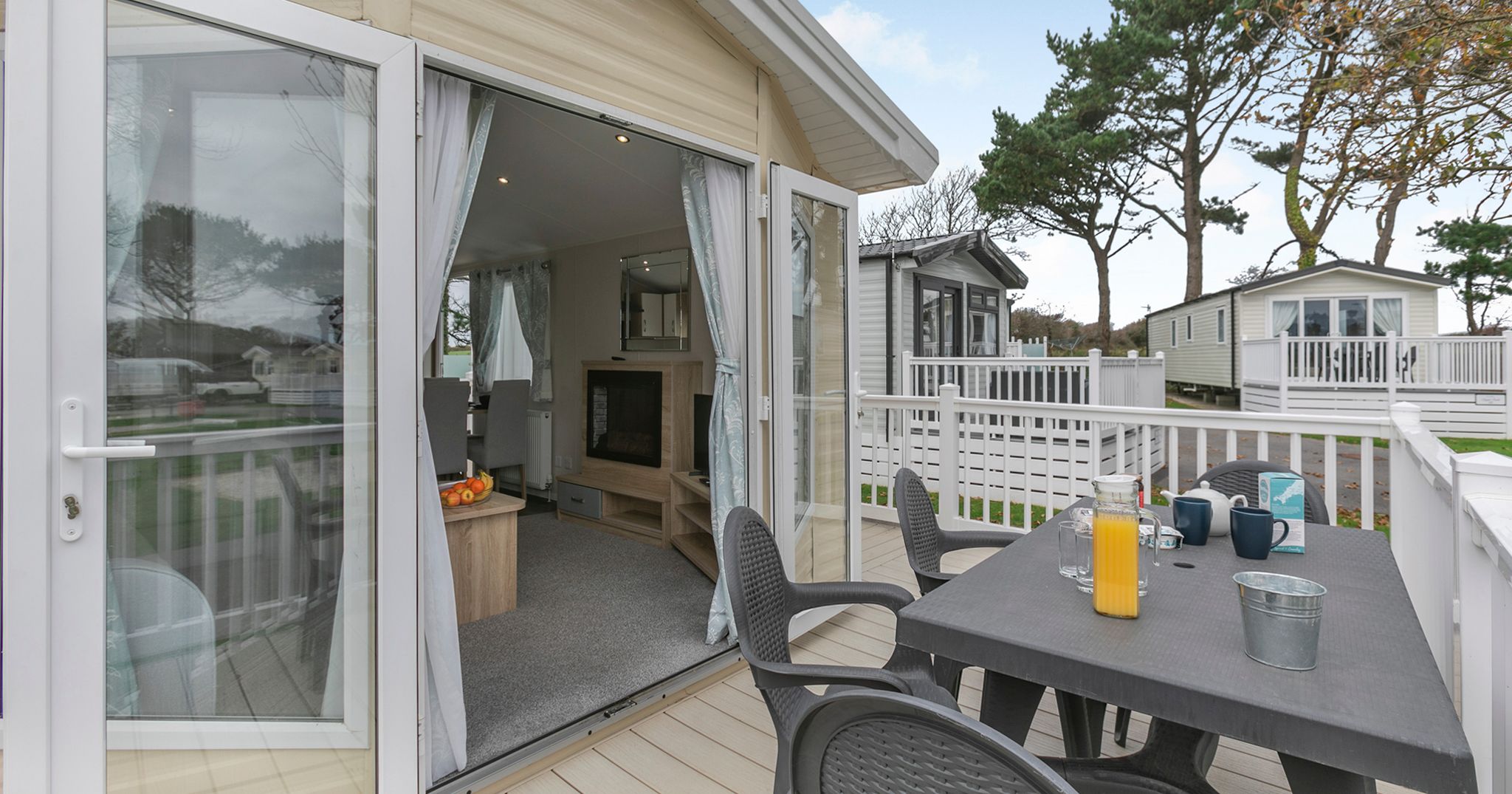 Modern mobile home with a deck featuring outdoor seating and a table set for breakfast, surrounded by similar units and trees.
