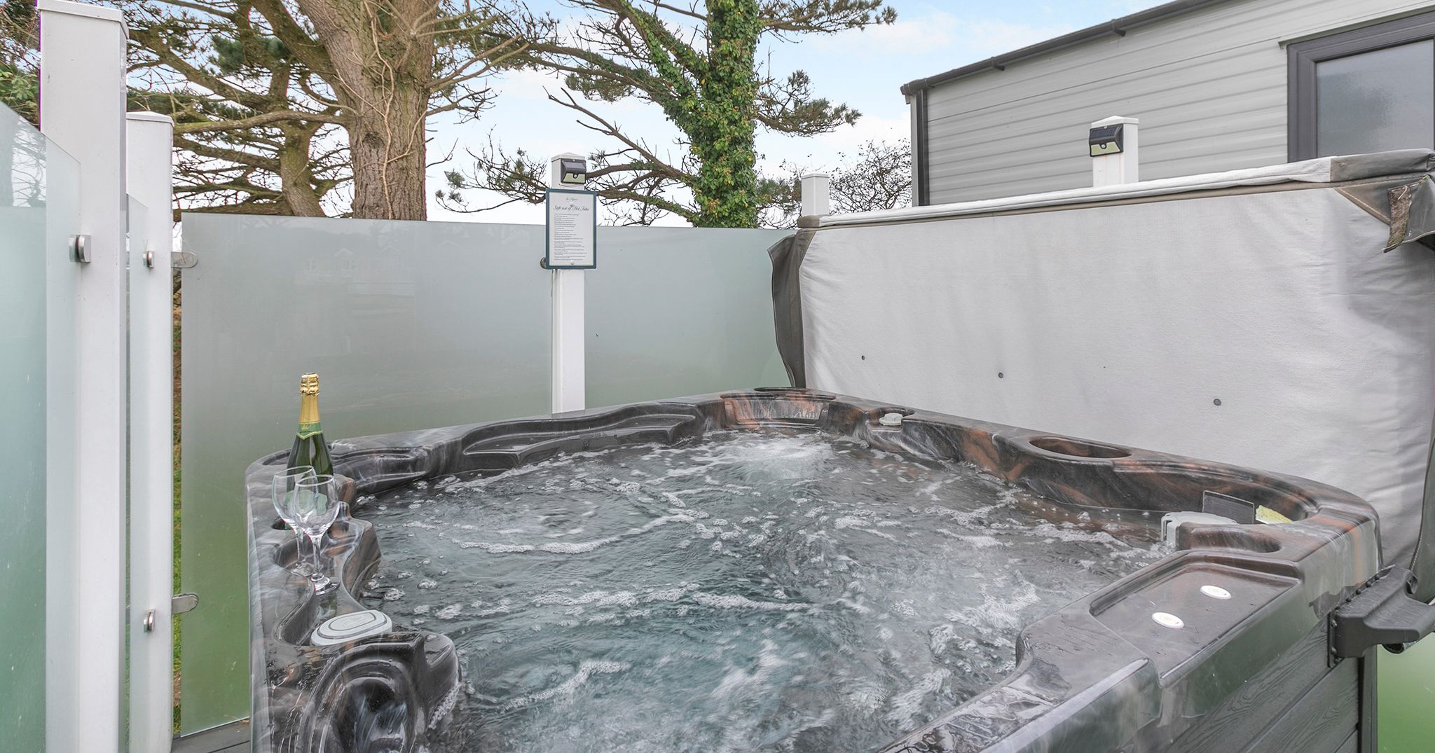 Outdoor hot tub with bubbling water beside a privacy screen and trees, with champagne and glasses on the edge.