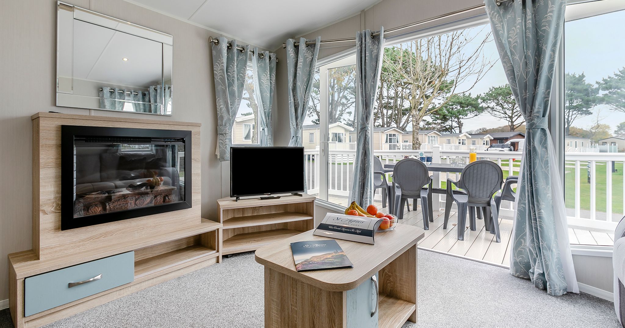 Modern living room with fireplace, TV, coffee table and large windows overlooking a patio with outdoor seating.