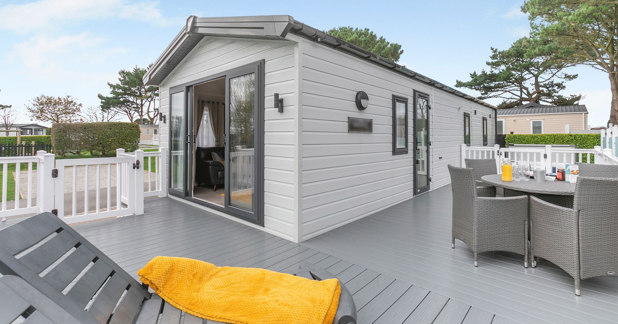 Modern mobile home with a spacious gray deck, outdoor seating, and lounge chairs with yellow blankets.