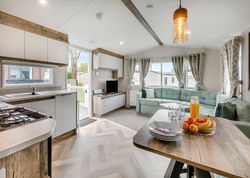 Modern open-plan kitchen and living room in a mobile home with large windows, a sectional sofa, and a dining table set with fruit and juice.