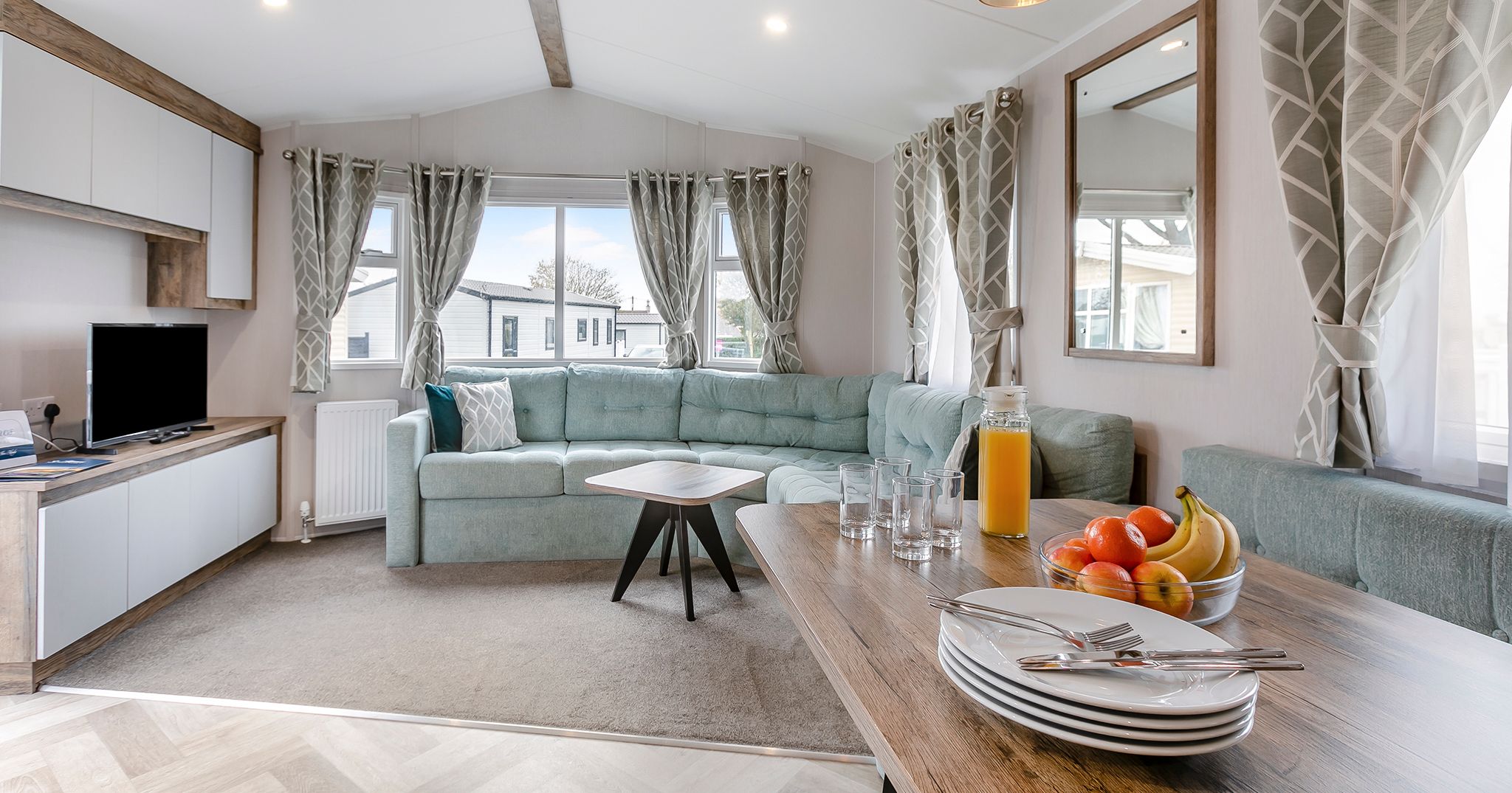 Modern living room and dining area inside a mobile home with a large sofa, dining table set with plates, cutlery, glassware, a jug of orange juice, and a bowl of fruit.
