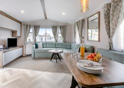 Modern living room and dining area inside a mobile home with a large sofa, dining table set with plates, cutlery, glassware, a jug of orange juice, and a bowl of fruit.