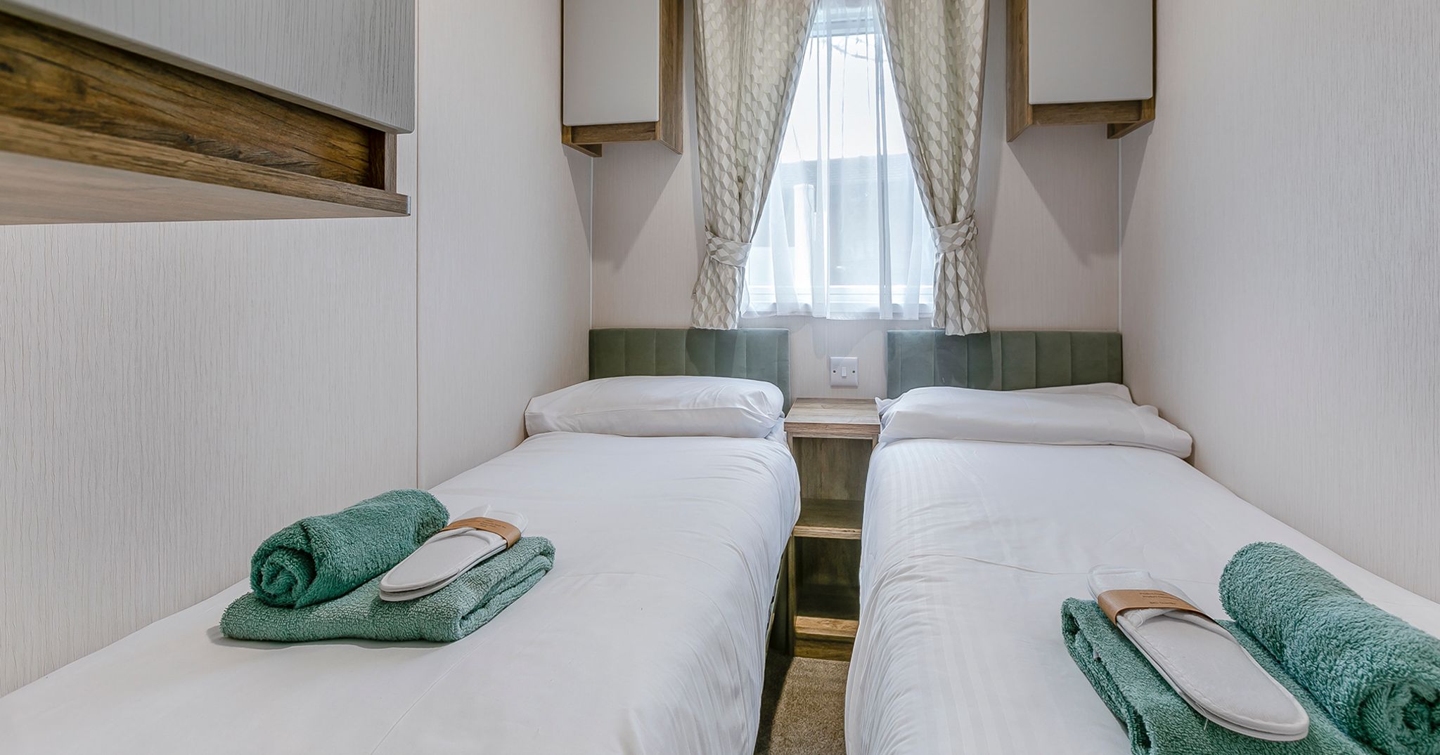 A small bedroom with two single beds, each with white bedding, green towels, and a pair of slippers at the foot of each bed.