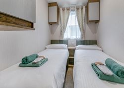 A small bedroom with two single beds, each with white bedding, green towels, and a pair of slippers at the foot of each bed.