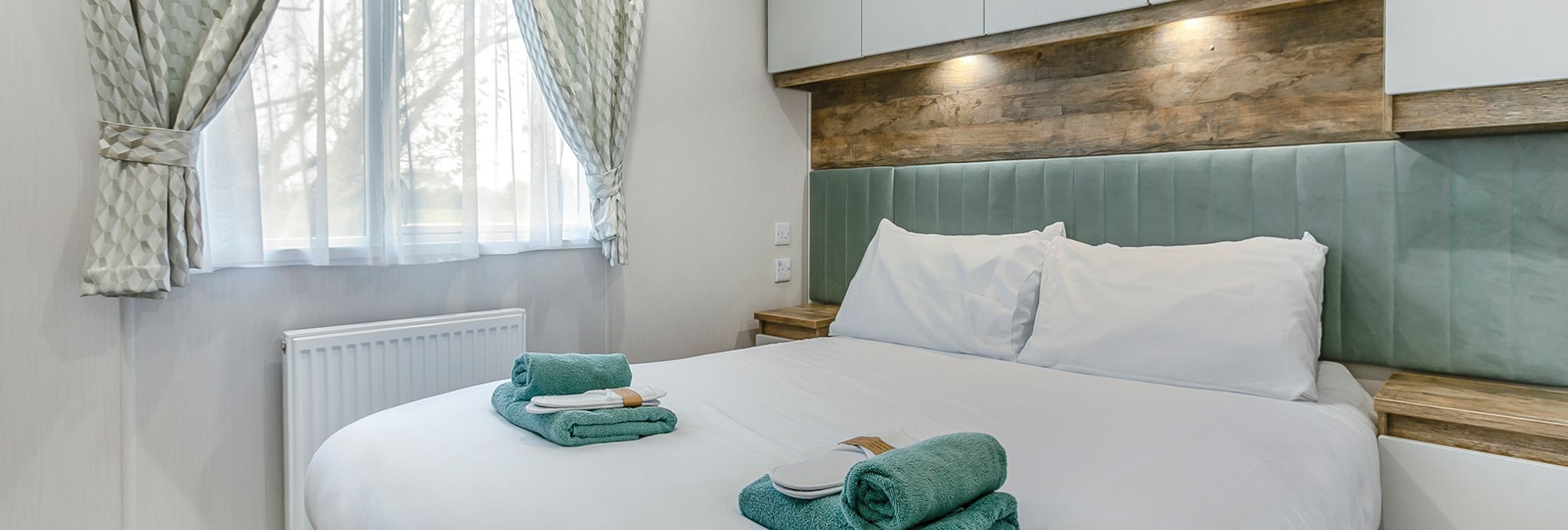 Modern bedroom with a large bed, two sets of folded green towels, and soft natural light coming through a window with curtains.