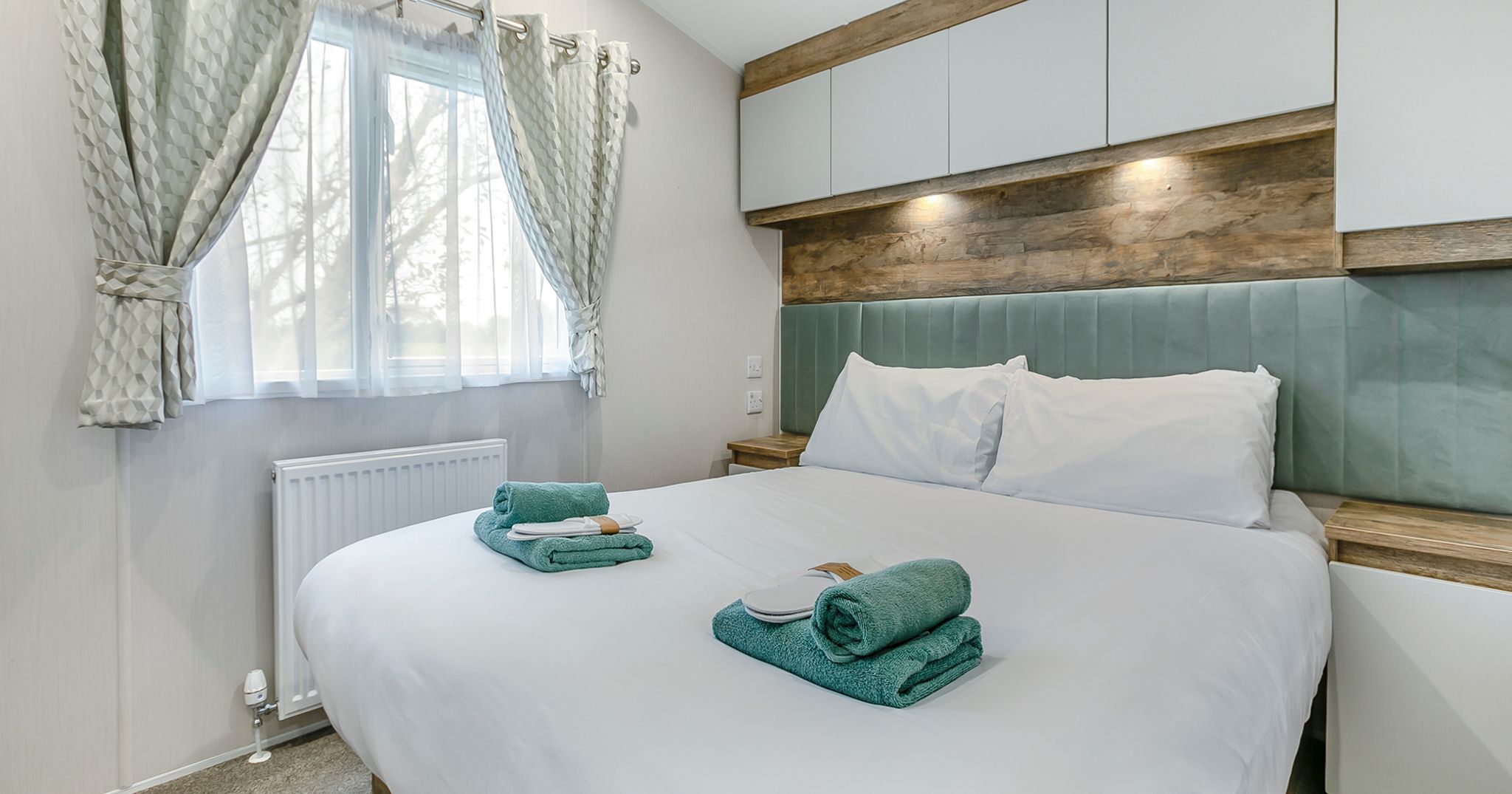 Modern bedroom with a large bed, two sets of folded green towels, and soft natural light coming through a window with curtains.