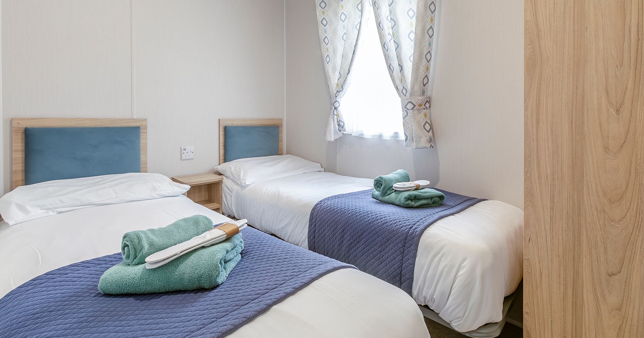 Two single beds with blue headboards and folded towels in a bright room with a window and patterned curtains.