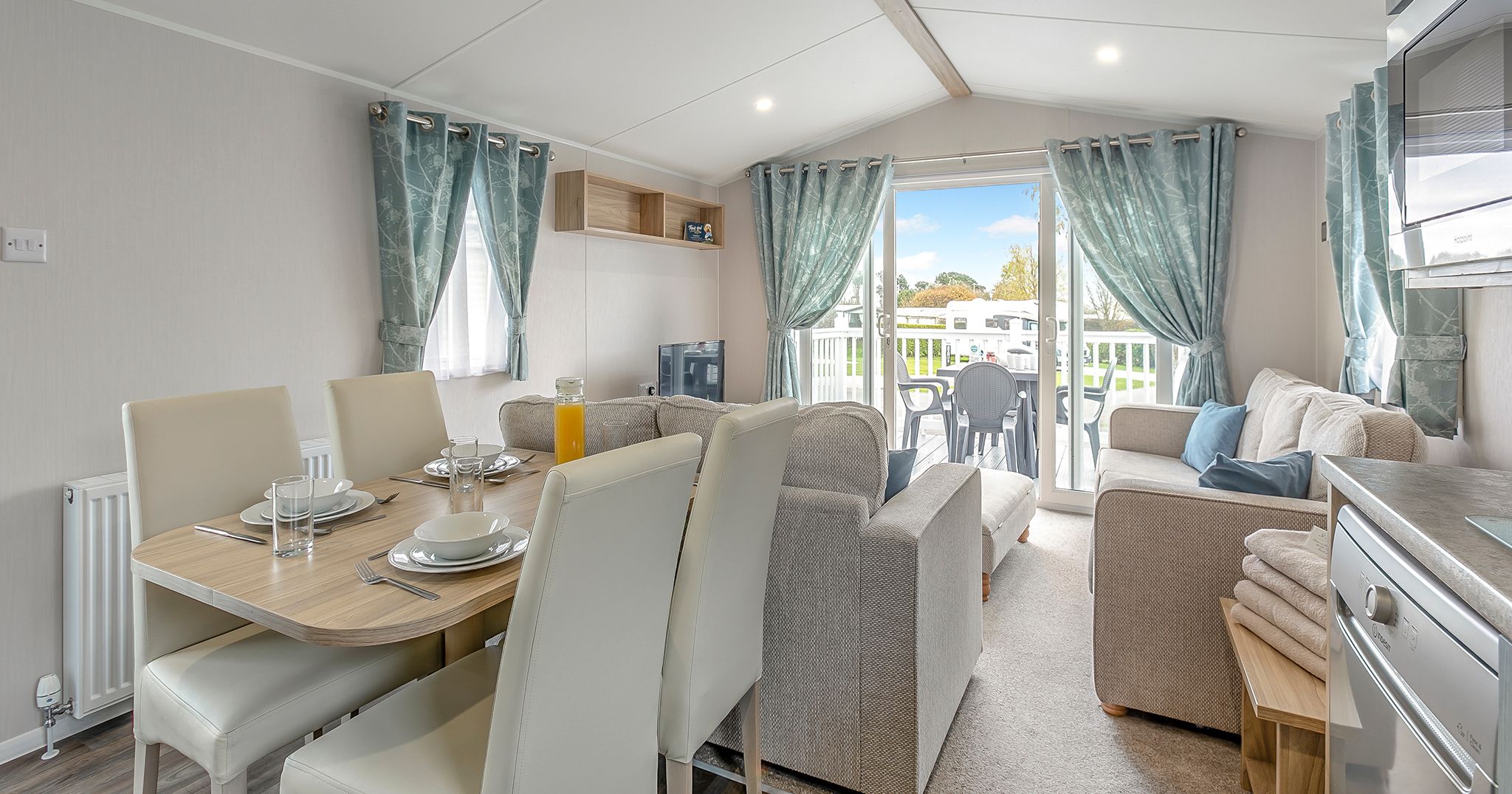 Modern living and dining area inside a caravan with large windows and patio doors opening to an outdoor deck.
