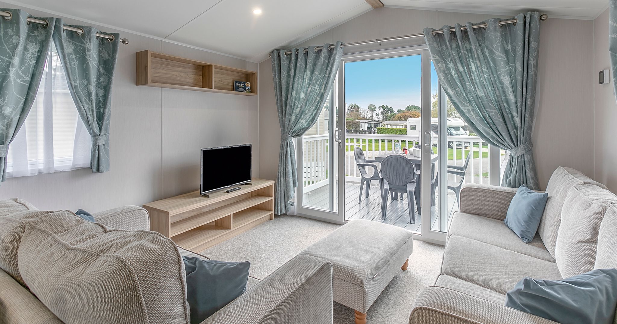 Modern living room with light-colored sofas, TV, and sliding glass doors opening to a deck with patio furniture.