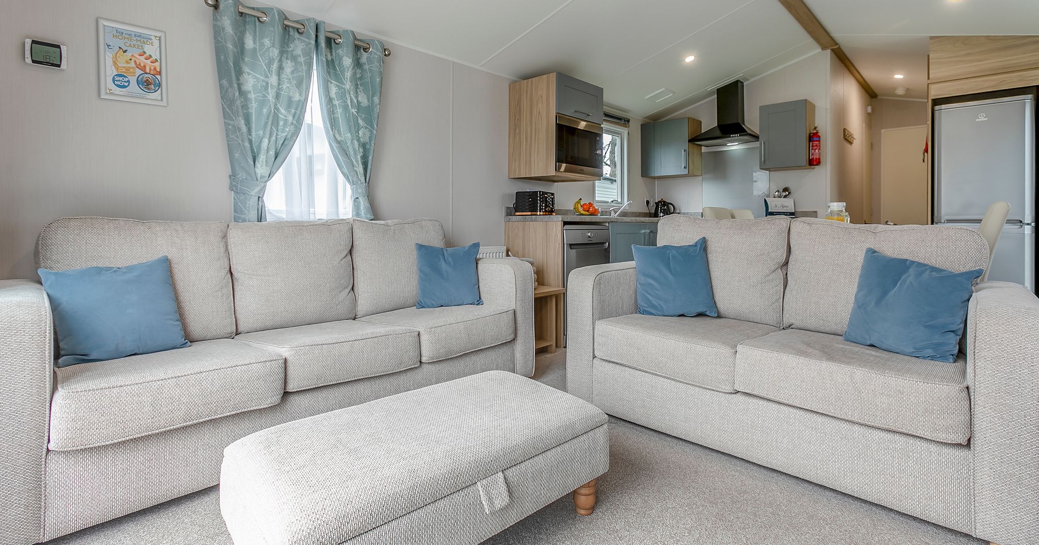 Modern living room with two beige sofas, blue cushions, and a matching ottoman in front of a kitchen area.