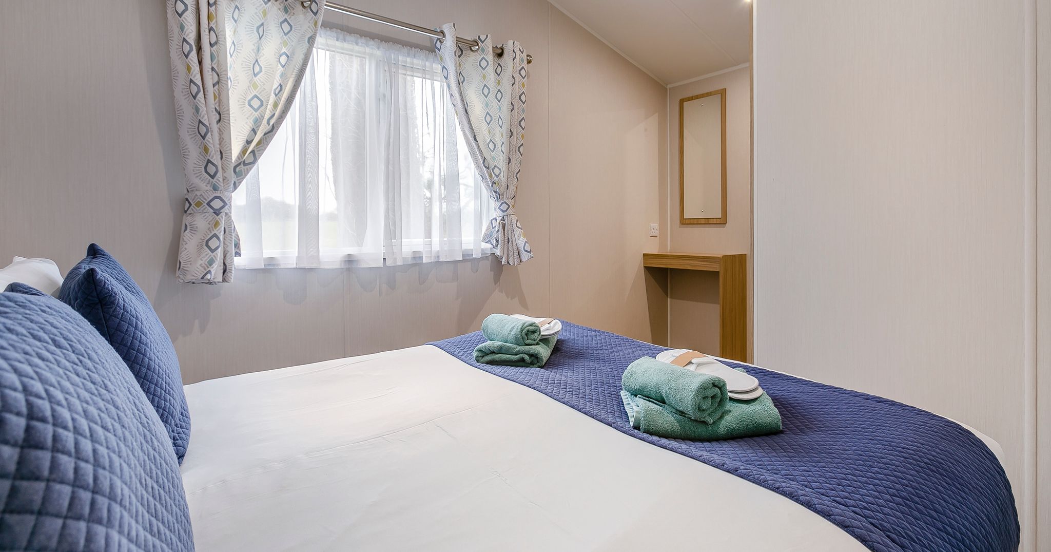 Bright bedroom with double bed, blue pillows and runner, two sets of towels, and a window with patterned curtains.