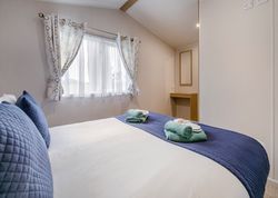 Bright bedroom with double bed, blue pillows and runner, two sets of towels, and a window with patterned curtains.