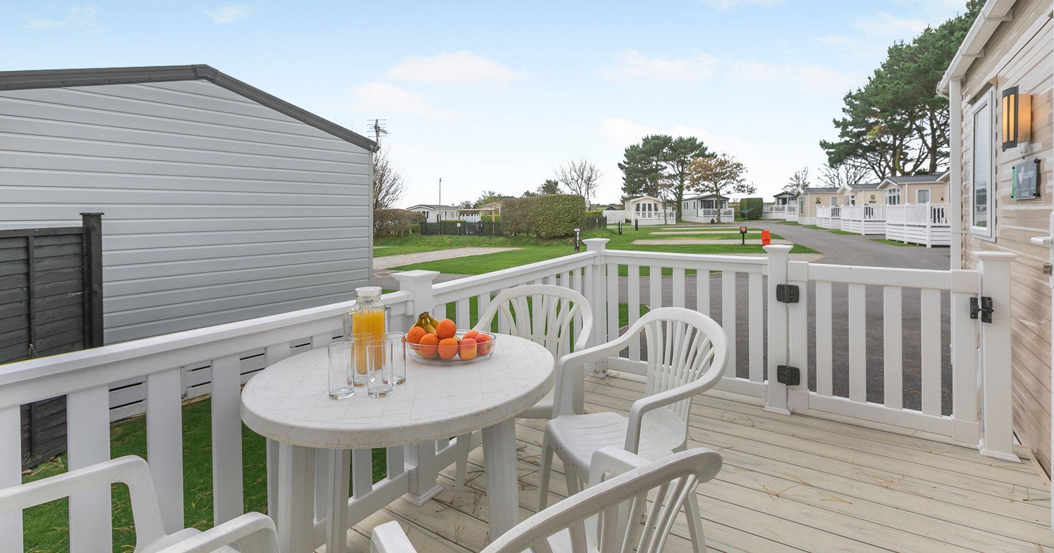 Outdoor patio area with white plastic chairs and table, fruit bowl and orange juice on table, and view of mobile homes.