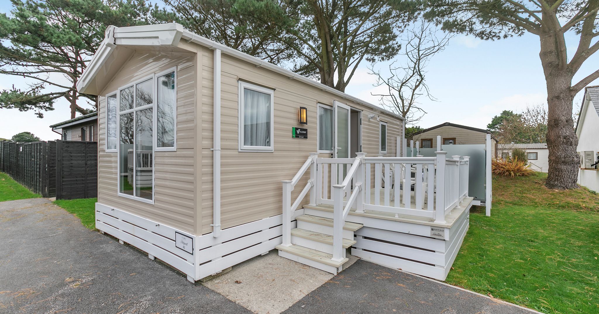 Modern beige mobile home with white porch and steps in a green, tree-filled park.