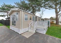 Modern beige mobile home with white porch and steps in a green, tree-filled park.