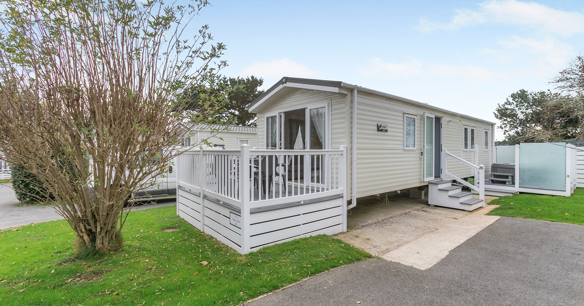 Modern mobile home with a white railing deck and steps, surrounded by greenery.