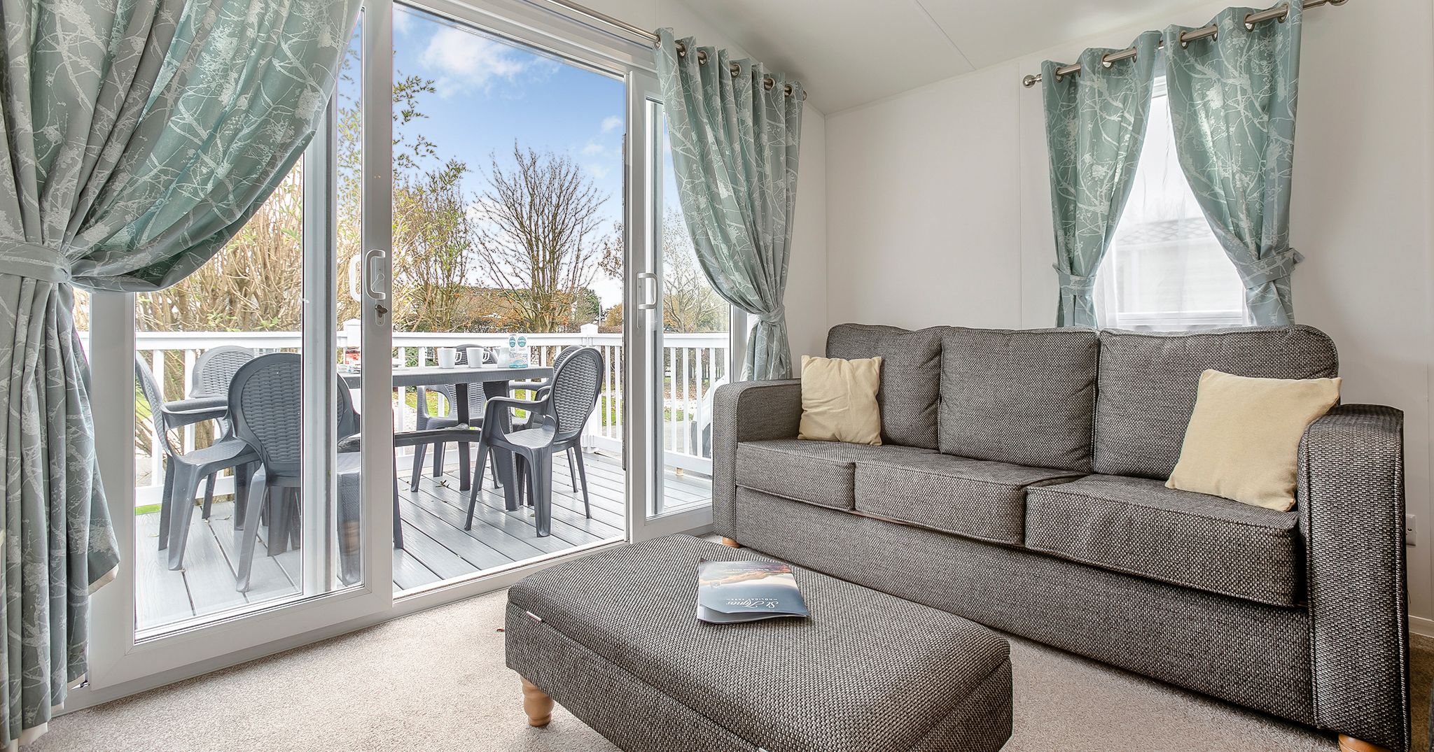 Modern living room with grey sofa, ottoman, and sliding glass doors leading to a patio with outdoor chairs and a table.