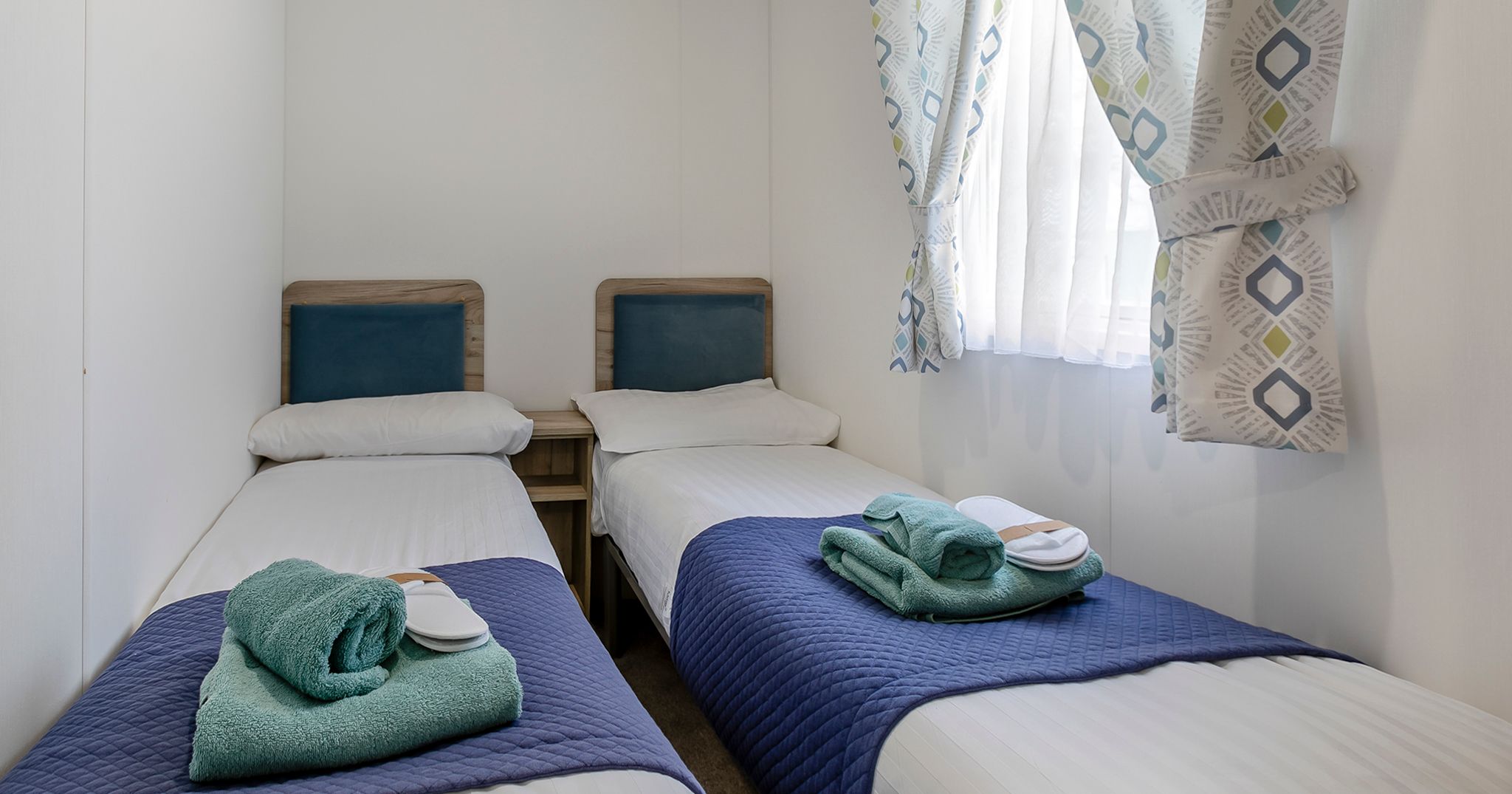 A small bedroom with two single beds, blue bed runners, and folded towels on each bed.