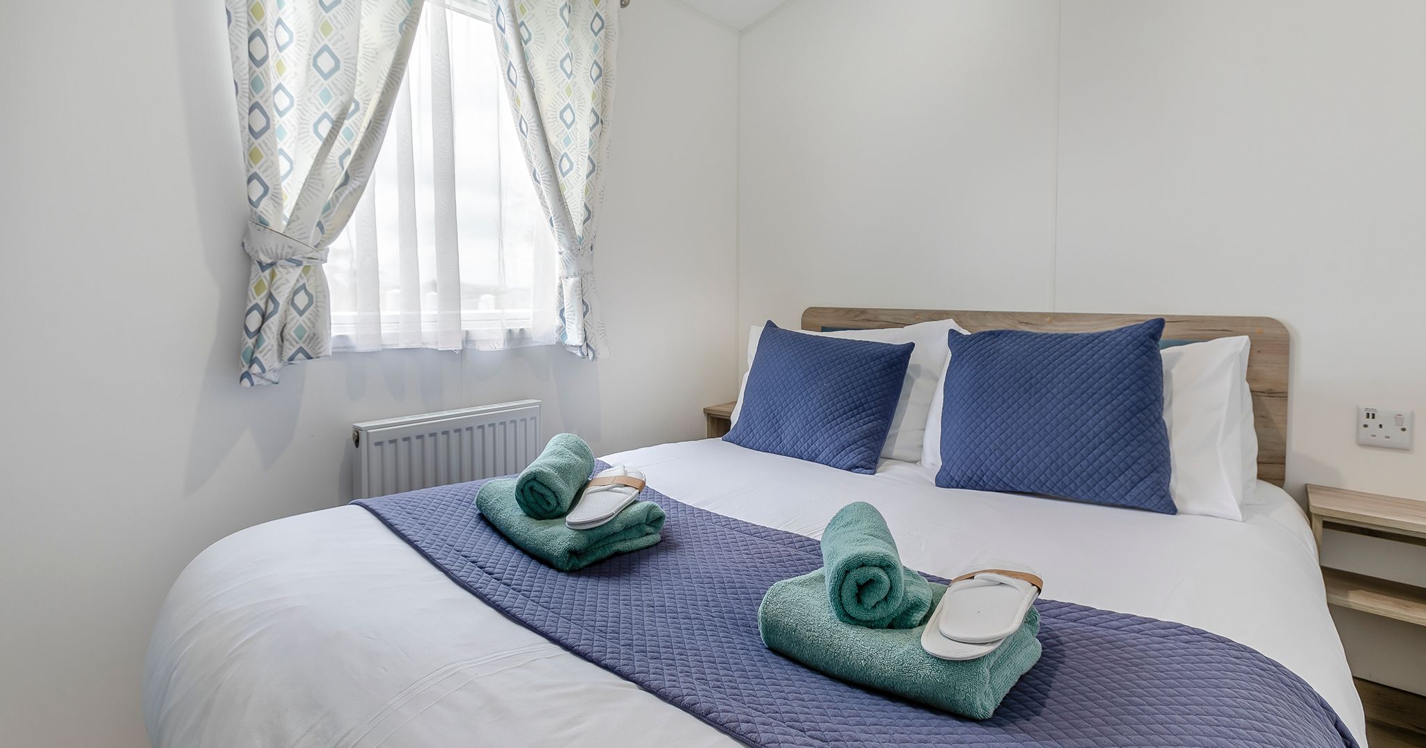 Neatly made bed with blue pillows, rolled towels, and slippers in a bright, simple bedroom.