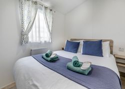 Neatly made bed with blue pillows, rolled towels, and slippers in a bright, simple bedroom.