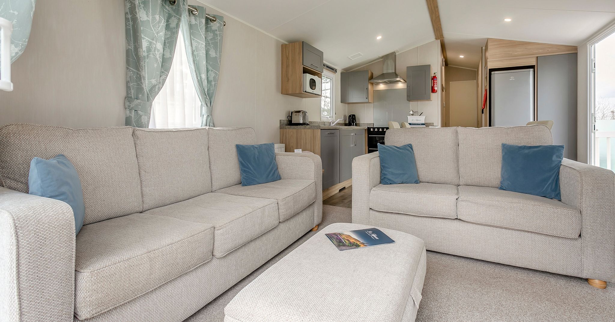 Modern living room with beige sofas, blue pillows, and a view into the kitchen.