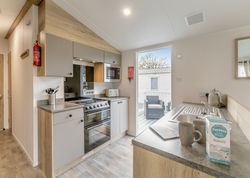 Modern kitchen in a mobile home with light wood and grey cabinets, stainless steel appliances, and outdoor seating visible through a glass door.