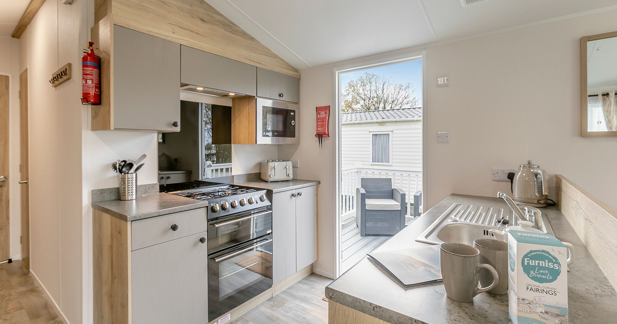 Modern kitchen in a mobile home with light wood and grey cabinets, stainless steel appliances, and outdoor seating visible through a glass door.