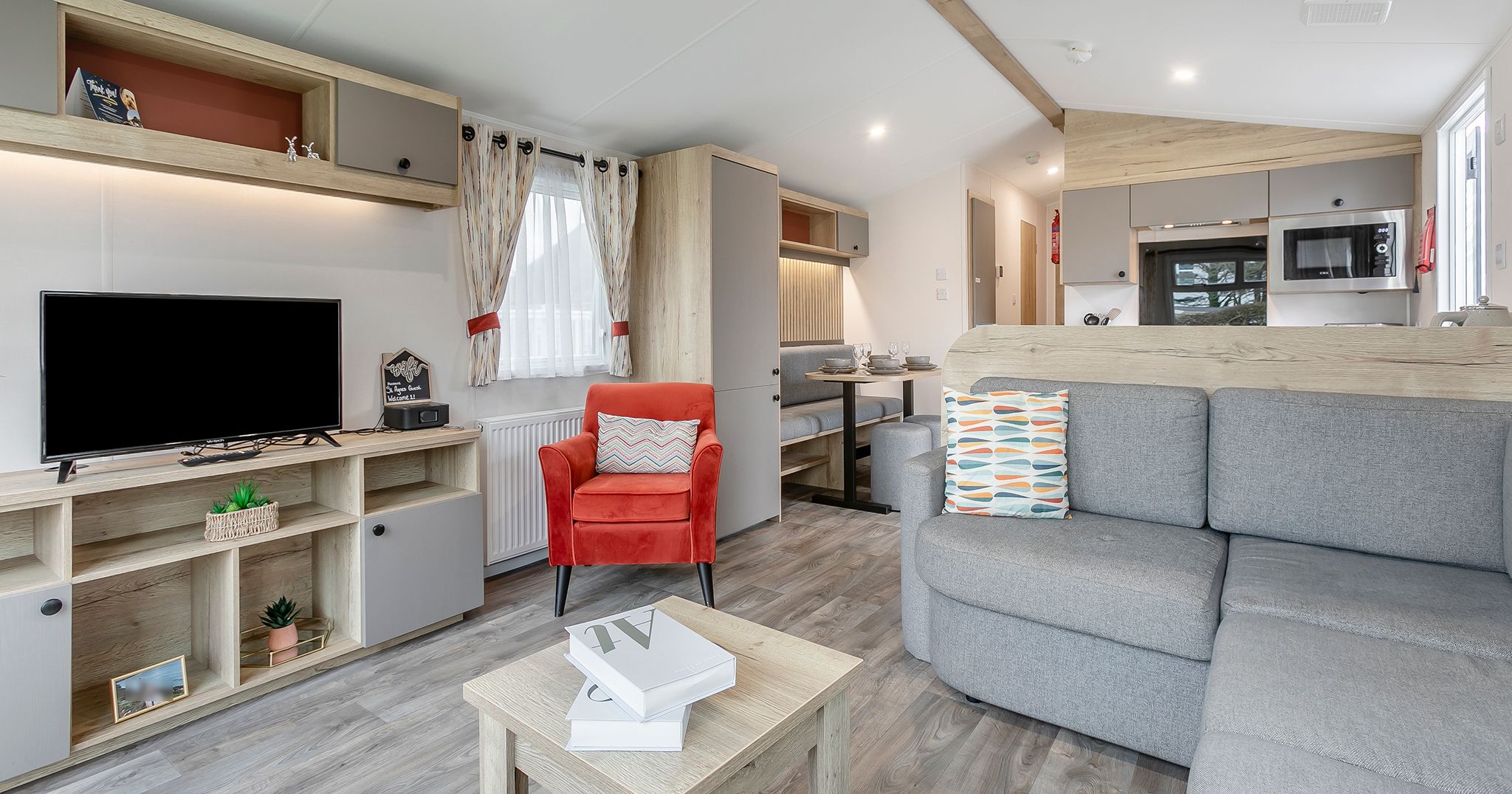 Modern open-plan living area with grey corner sofa, orange armchair, TV unit, and kitchen