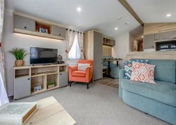 Modern, cozy living room in a mobile home featuring a TV, orange armchair, light blue sofa with patterned cushions, and a small dining area.