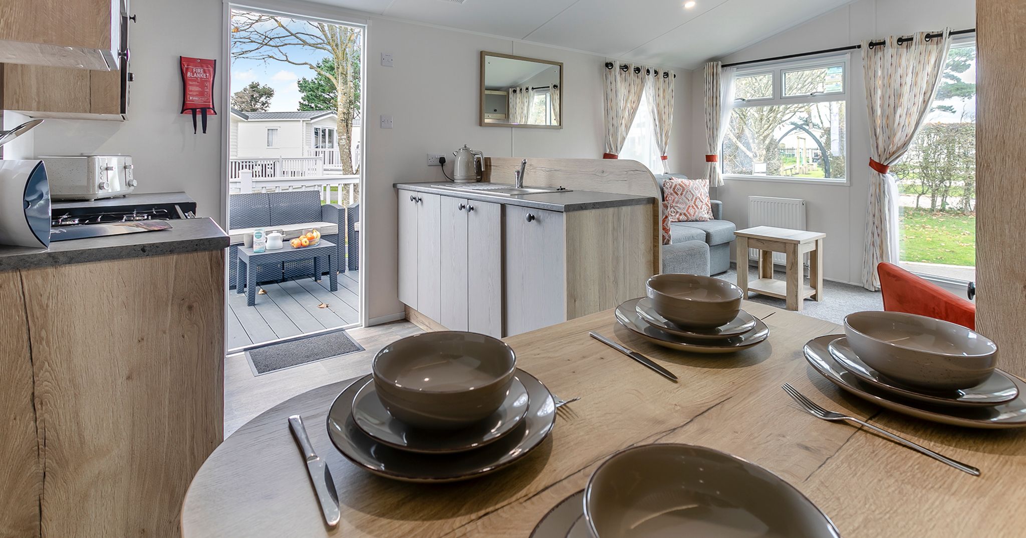 Modern open-plan kitchen and living area in a mobile home with table set for four, large windows, and an outdoor seating area visible through an open door.