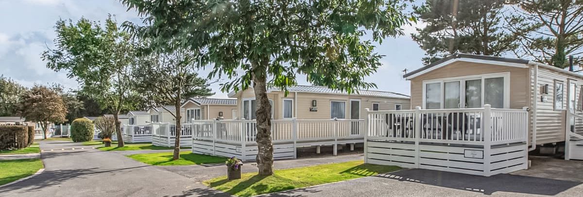 Row of modern mobile homes with white decking under trees in a holiday park.