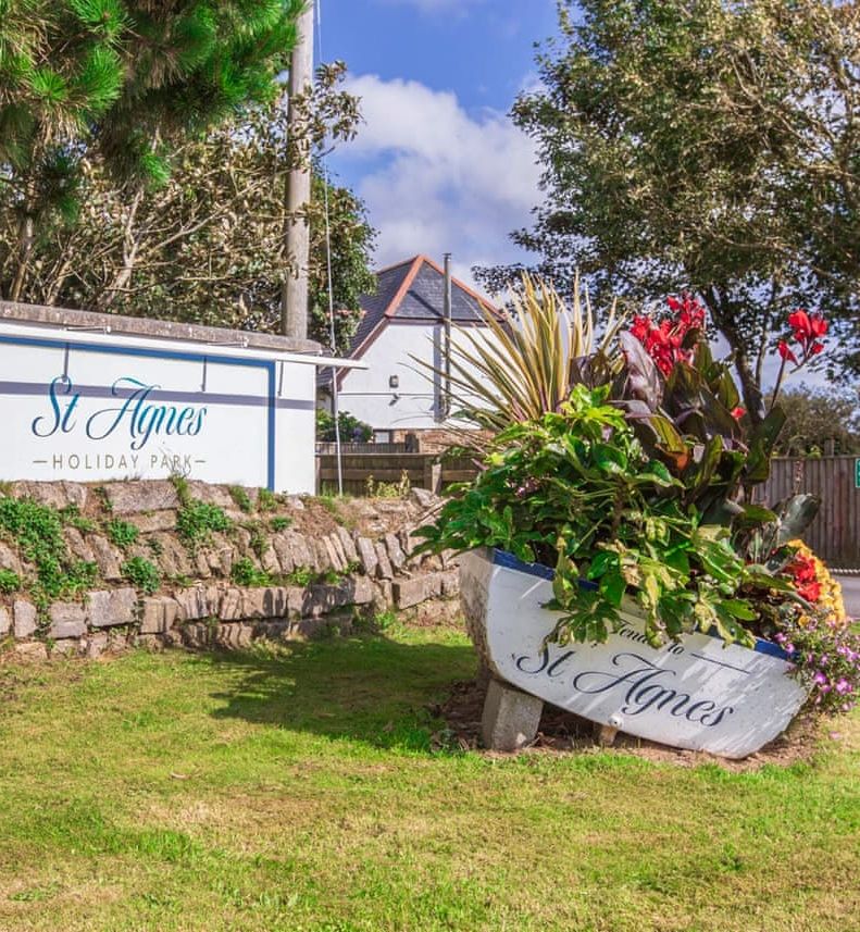 Entrance sign for St Agnes Holiday Park with a decorative boat planter filled with vibrant flowers.