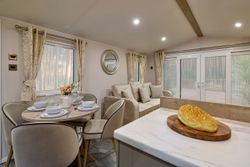 Modern living and dining space with a round table set for four, beige sofa, large windows, and a loaf of bread on a kitchen counter.