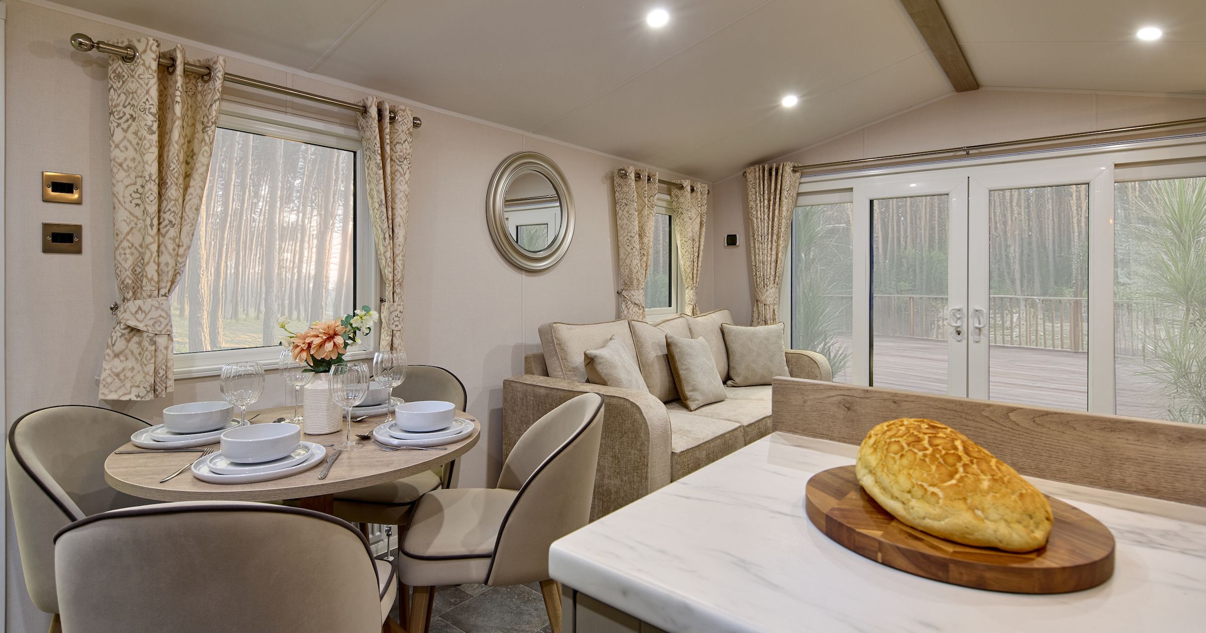 Modern living and dining space with a round table set for four, beige sofa, large windows, and a loaf of bread on a kitchen counter.