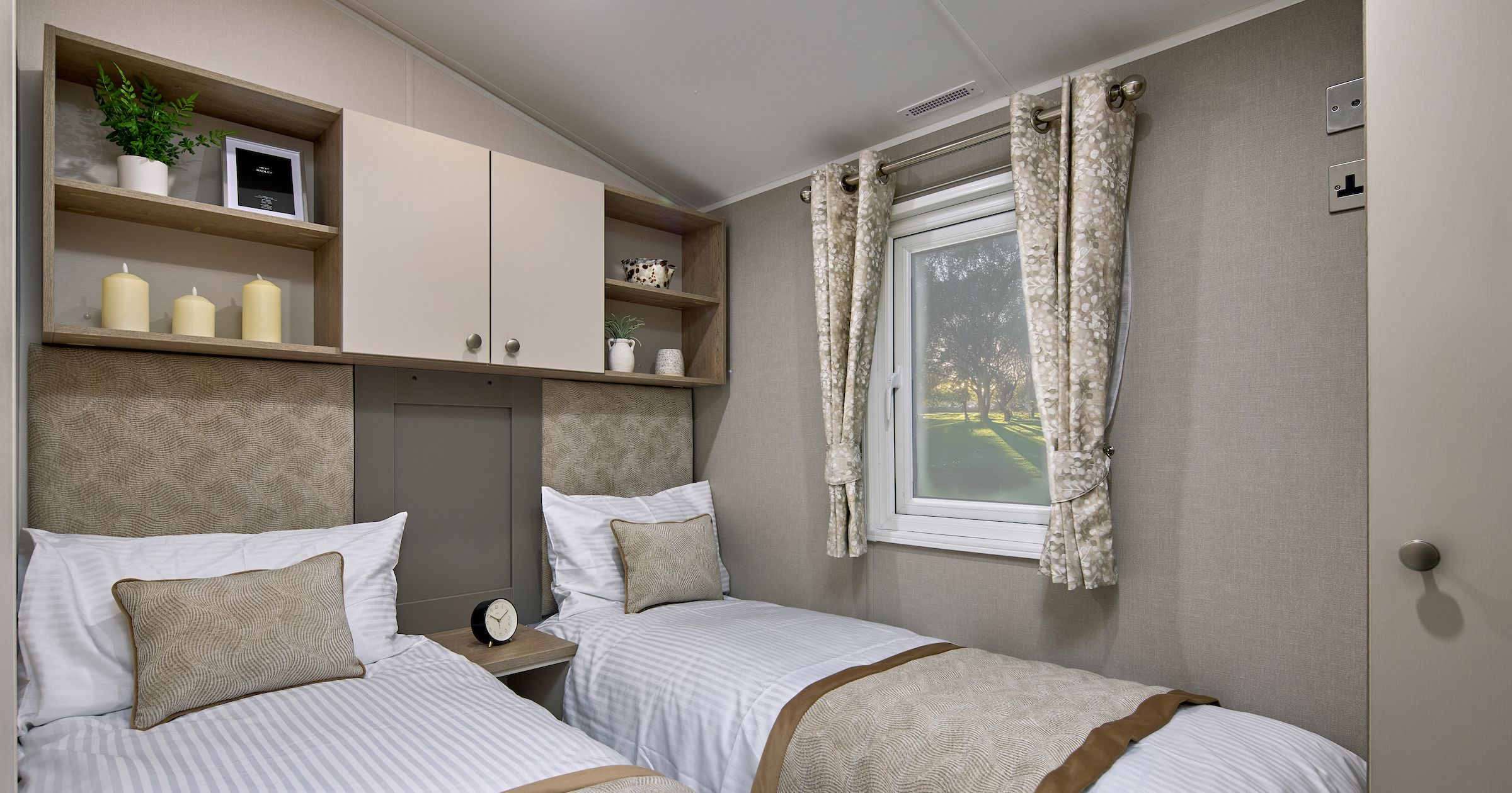 A neatly arranged small bedroom with two single beds, beige decor, a window with curtains, and built-in shelving above the beds.