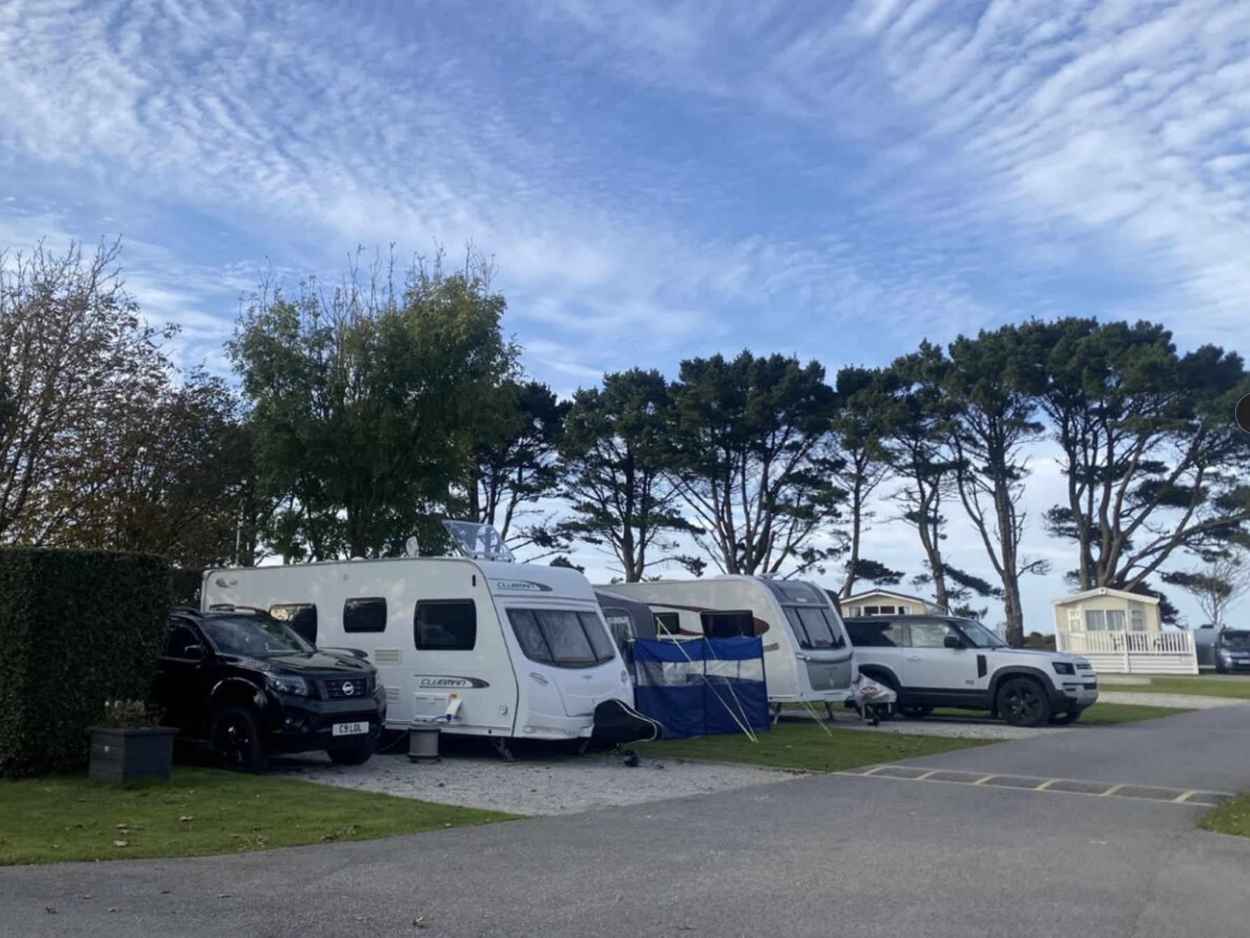 Caravans and cars parked at a campsite with trees and a blue sky in the background.