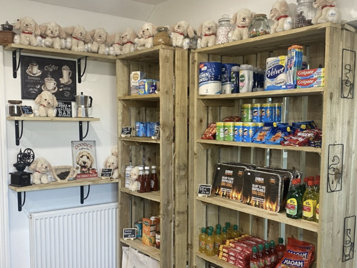 Small shop corner with wooden shelves holding snacks, drinks, toiletries, groceries, and plush toy dogs.