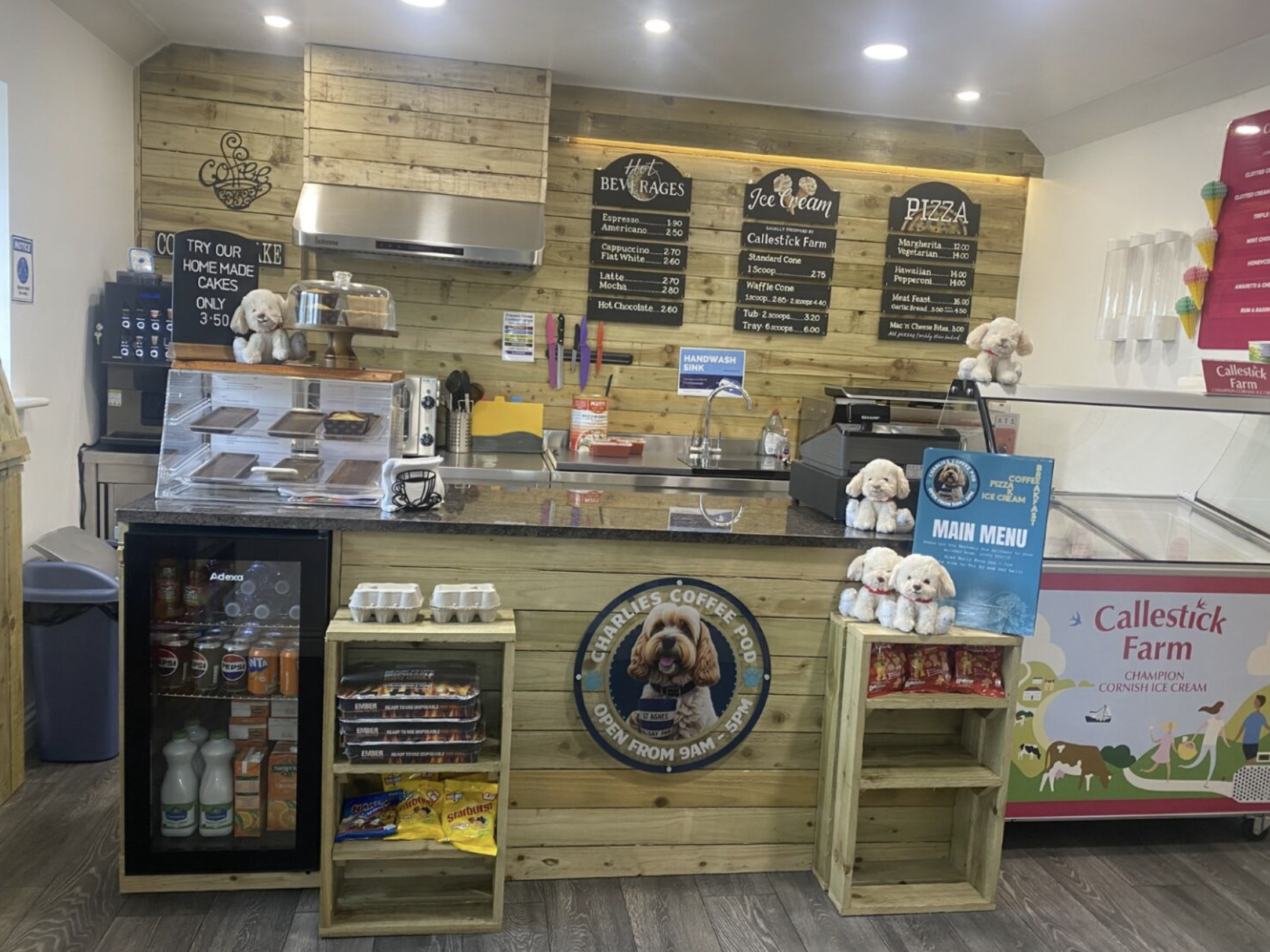 A cozy coffee shop counter with wooden decor, stuffed dog toys, snacks, and a menu featuring cakes, pizza, and ice cream.