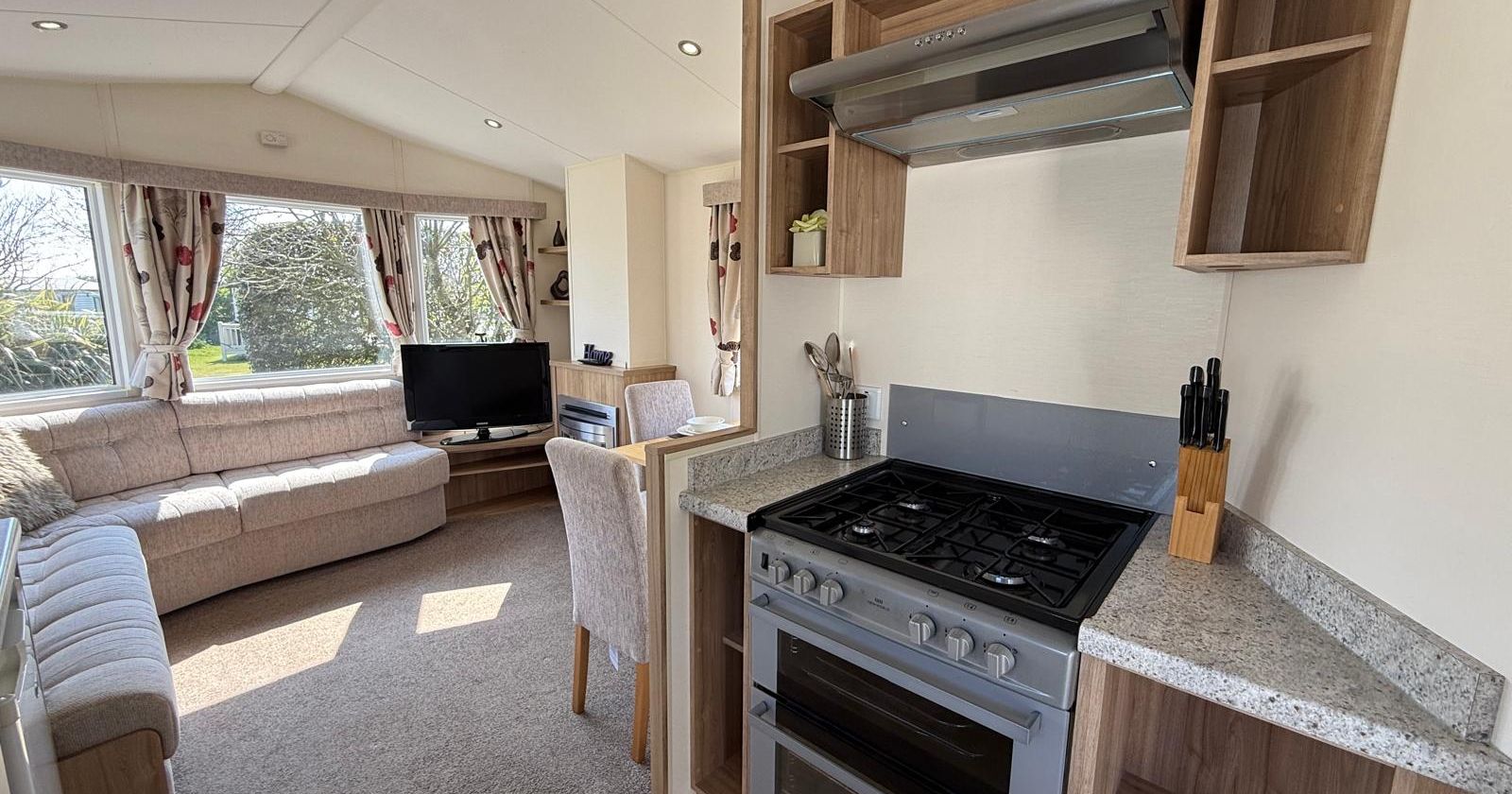 Modern caravan interior with a kitchen and living area featuring a stove, TV, and large corner sofa.
