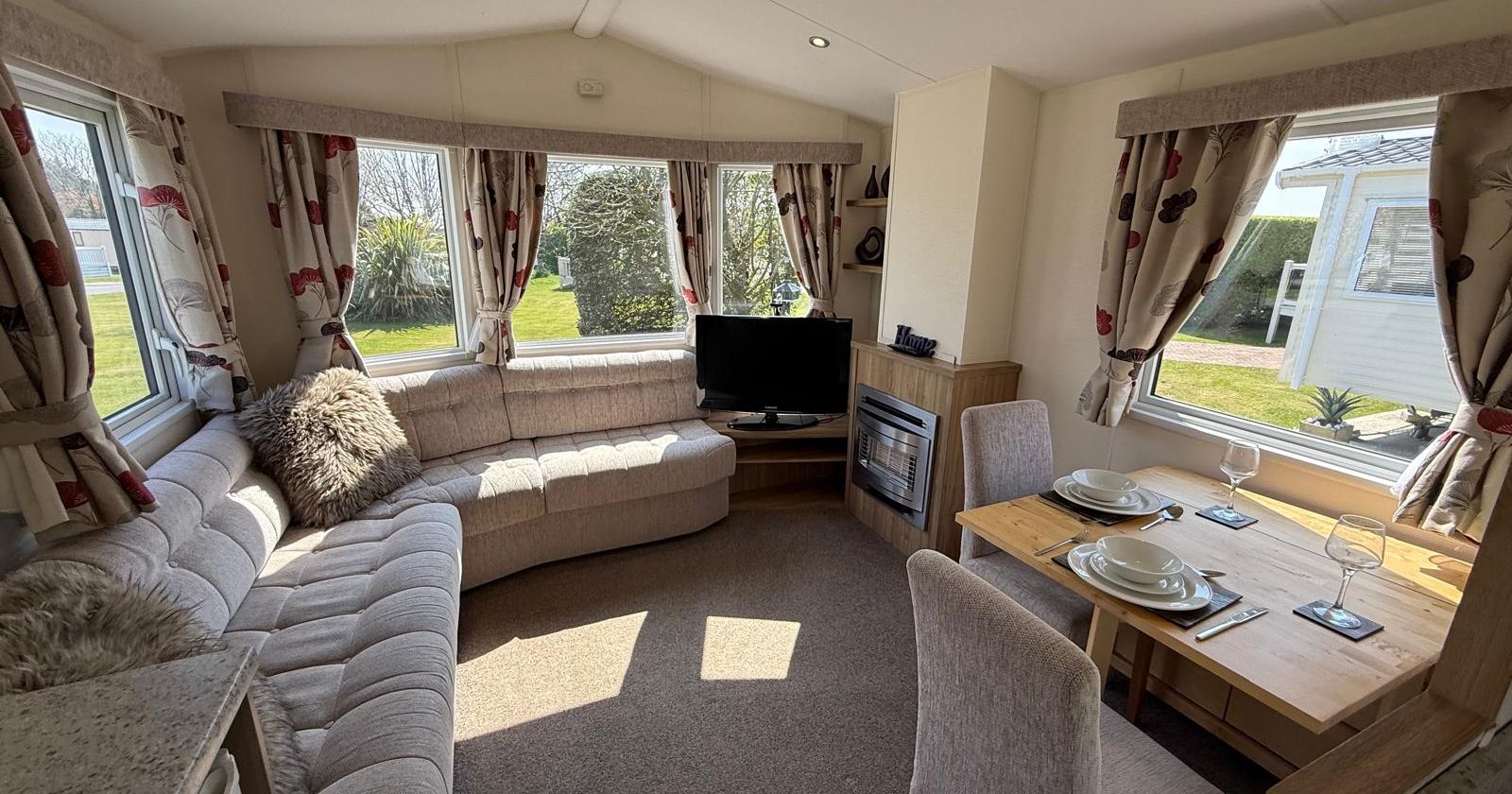 Bright and cozy living area and dining space inside a mobile home with windows overlooking green lawn.