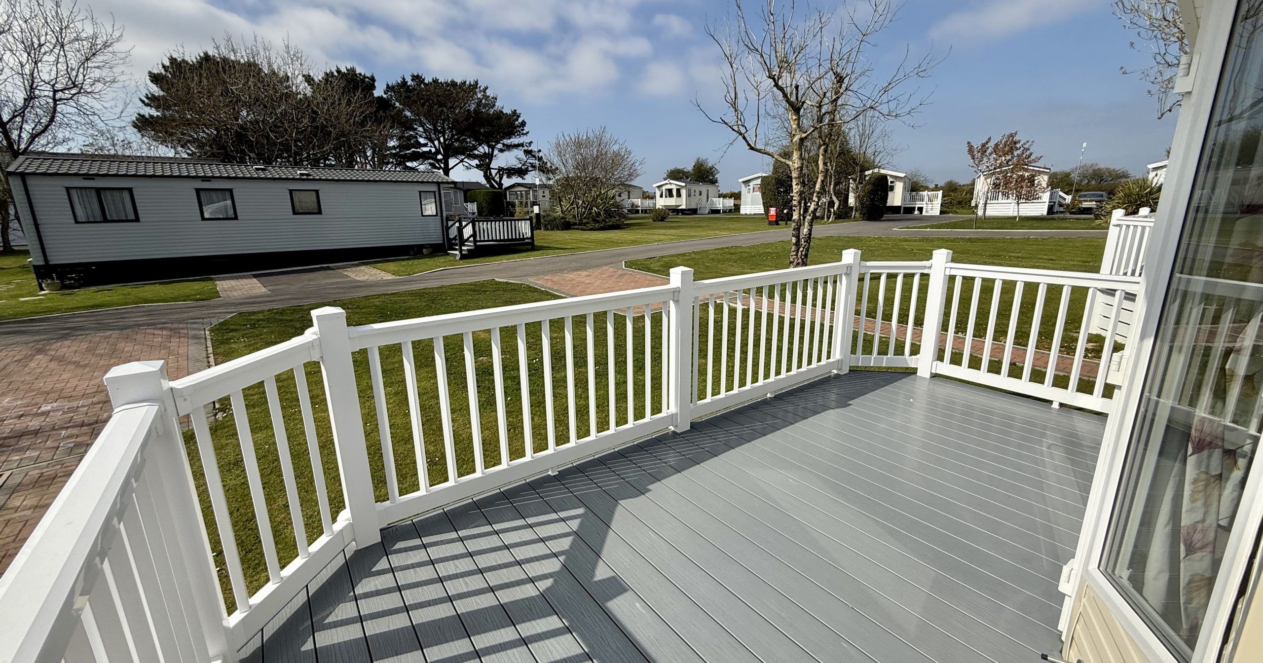 View from a white fenced deck overlooking a row of mobile homes on a grassy lot under a partly cloudy sky.