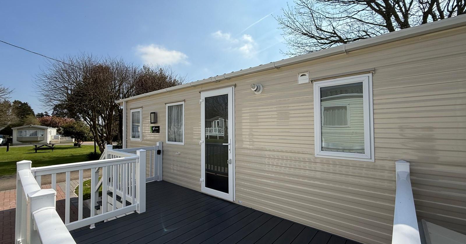 Exterior of a beige mobile home with white trim, porch, and surrounding grassy area under a clear sky