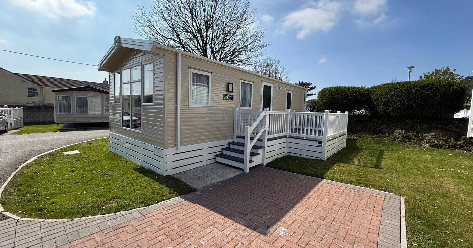 Modern beige mobile home with white fencing, steps, and a paved driveway on a sunny day