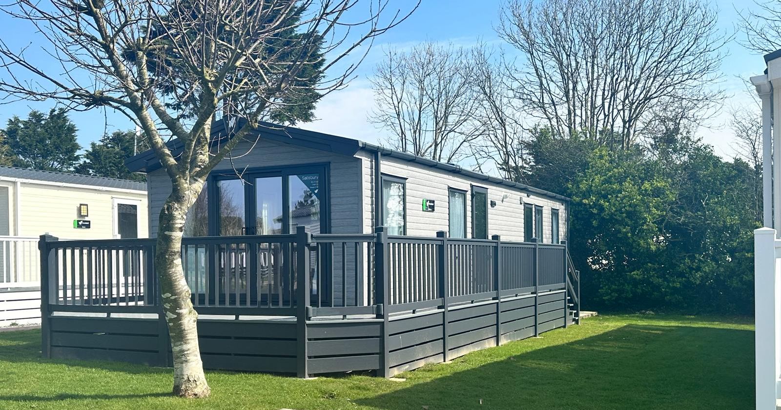 Modern grey holiday lodge with raised decking and railings beside a lawn and bare tree