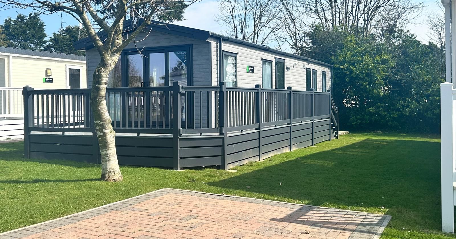 Modern grey holiday lodge with raised decking and railings beside a lawn and bare tree