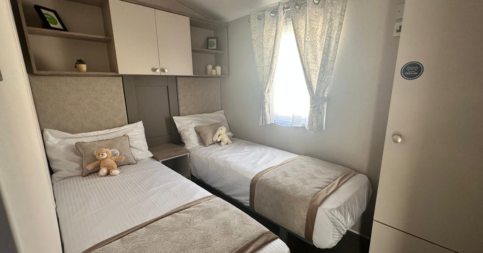 Compact twin bedroom with two single beds, overhead cupboards and a window with patterned curtains