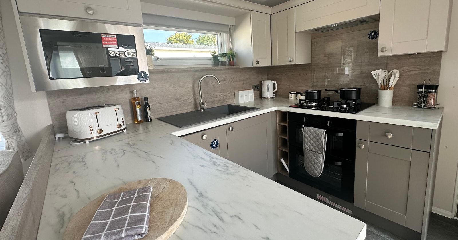 Modern L-shaped kitchen with marble-effect worktop, white and grey cabinets, sink under window, and gas hob with oven