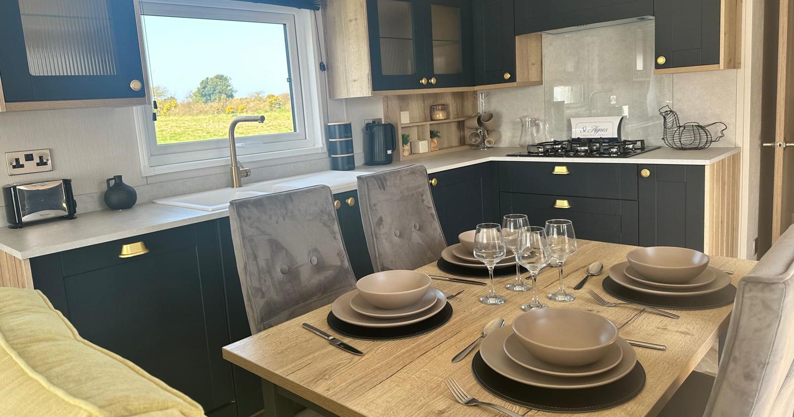 Modern caravan kitchen with black and wood cabinets, dining table set for four, and window overlooking countryside