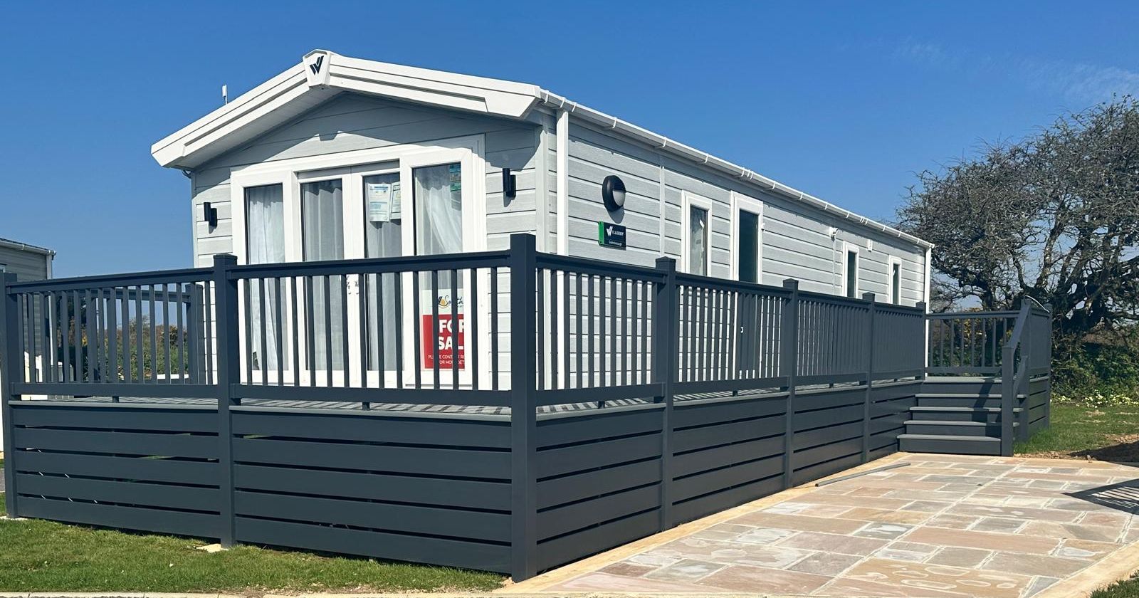 Light grey static caravan with a raised dark grey deck and railings under a clear blue sky